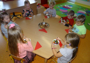 Grupa dzieci siedzi przy stole nakrytym serwetkami. Przy każdym dziecku ustawiona jest miseczka z sałatką owocową, którą jedzą dzieci.